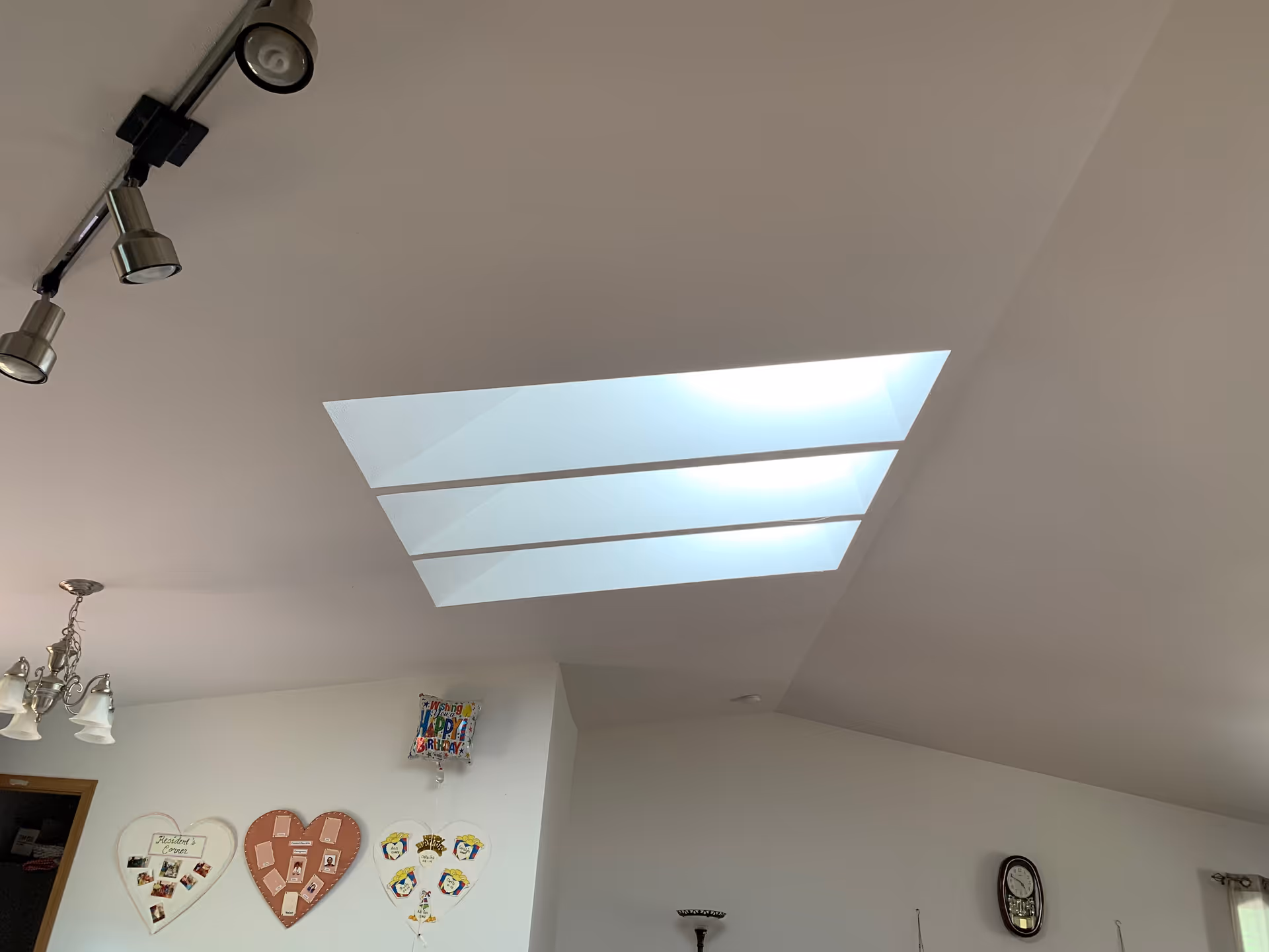 Interior view of a room ceiling featuring four rectangular skylights, track and chandelier lighting, and a wall with heart-shaped photo displays and a clock.