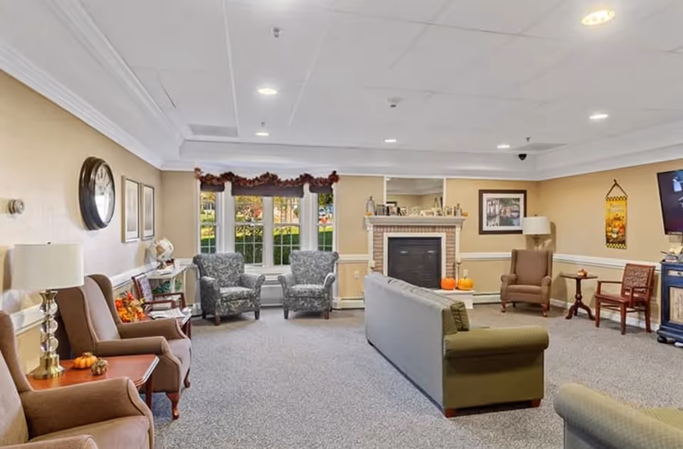 A cozy living room in a senior living facility with beige walls and carpeted floor. The room features a fireplace with decorative items on the mantel, two patterned armchairs near a window with fall-themed garland, several brown armchairs, a green sofa, side tables with lamps, and a wall-mounted TV. The room is decorated with autumn-themed accents including small pumpkins.