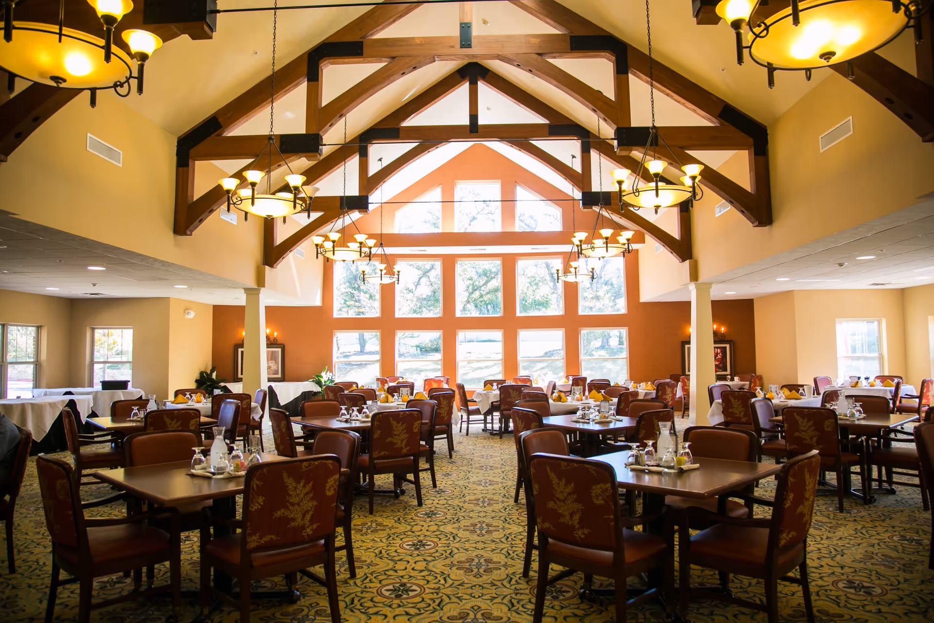 A spacious dining room with multiple tables and chairs arranged neatly. The room features large windows letting in natural light, high vaulted ceilings with wooden beams, and several chandeliers providing warm lighting. Tables are set with glassware and condiments, and the carpet has a decorative pattern.