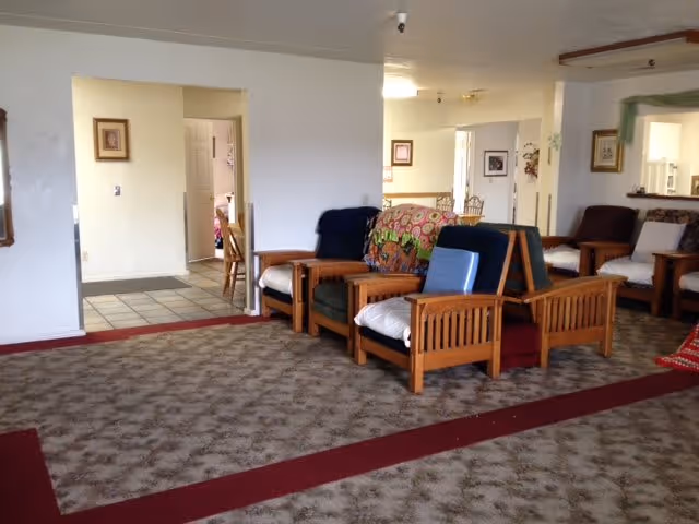 A living room area with several wooden armchairs arranged in two rows, some with cushions and blankets. The room has patterned carpet flooring with a red border and white walls. There are doorways leading to other rooms, including a glimpse of a dining area and a kitchen with white cabinets.