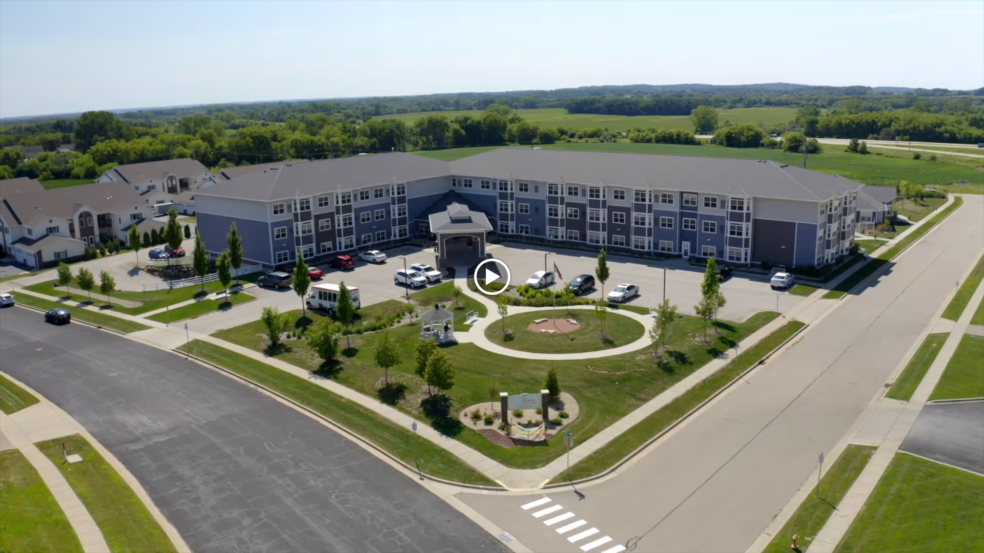 Aerial view of a three-story senior living building with a circular driveway, landscaped lawn, and parked cars.