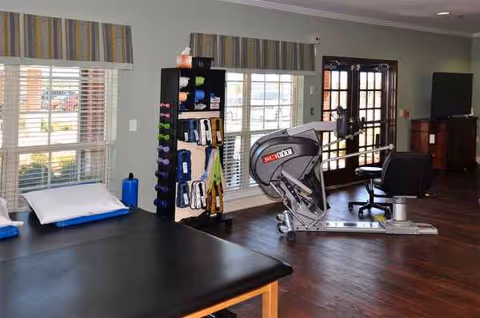 Indoor rehabilitation room with stationary exercise bike, black padded table, and shelving.