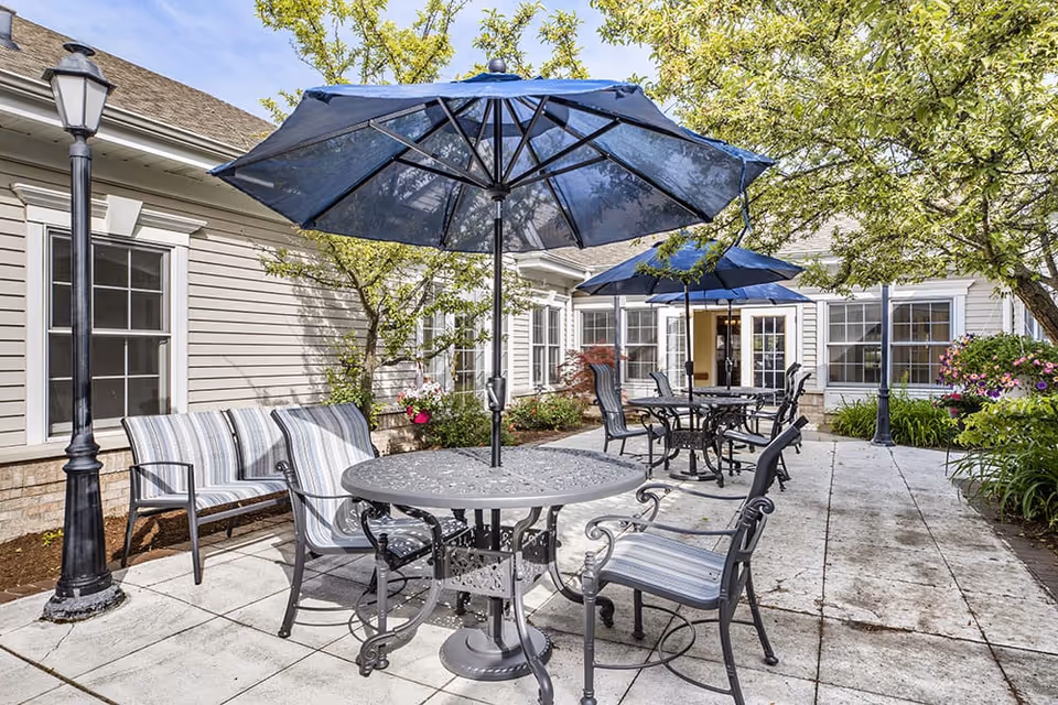 Outdoor patio area at Brookdale Troy featuring metal tables with blue umbrellas, striped cushioned chairs, a bench, lamp posts, and surrounding greenery including trees and flowering plants.