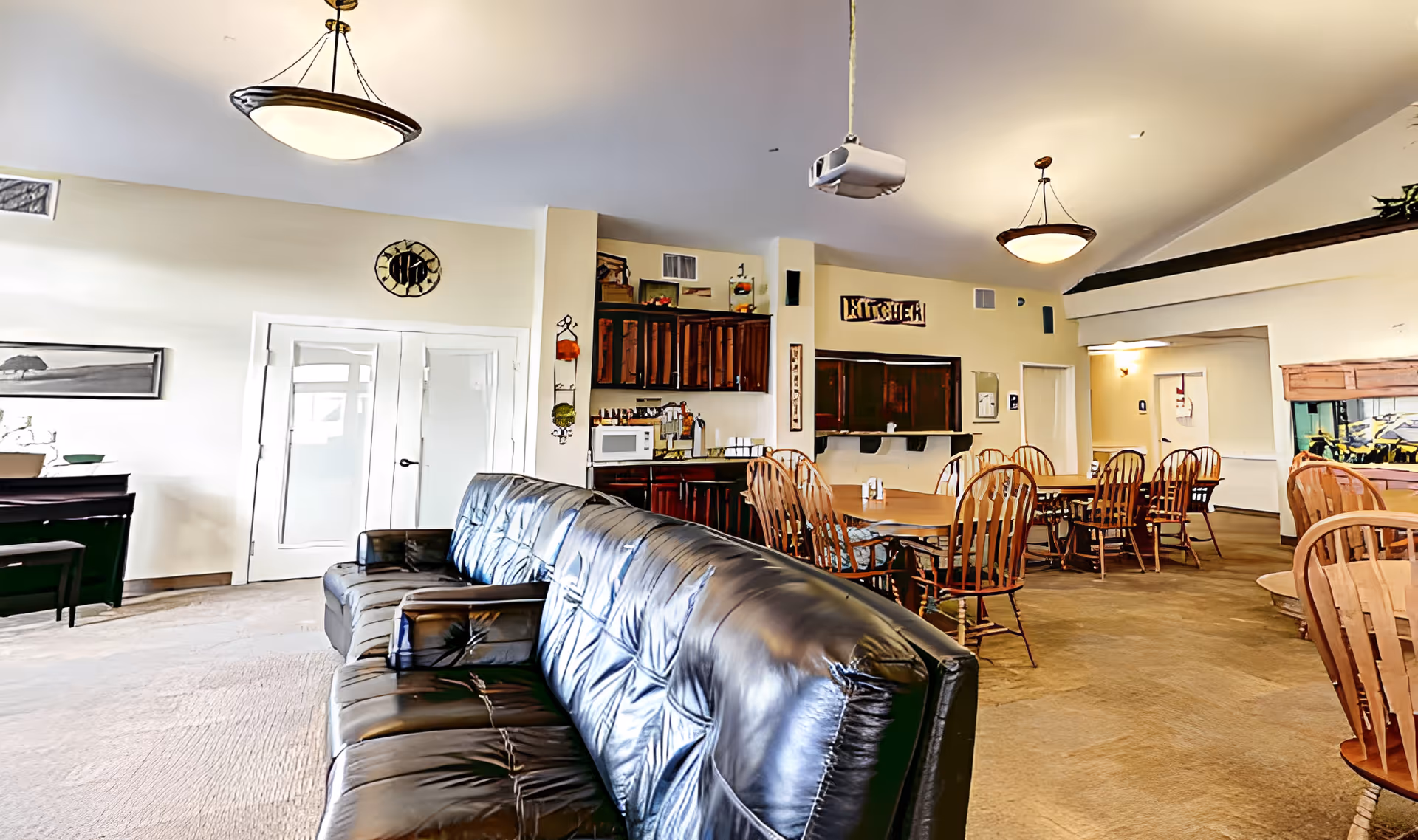 Interior view of a senior living facility common area featuring a black leather couch in the foreground, wooden dining tables with chairs, a small kitchen area with cabinets, a microwave, and a sign that reads 'KITCHEN'. The room is well-lit with ceiling lights and has a warm, inviting atmosphere.