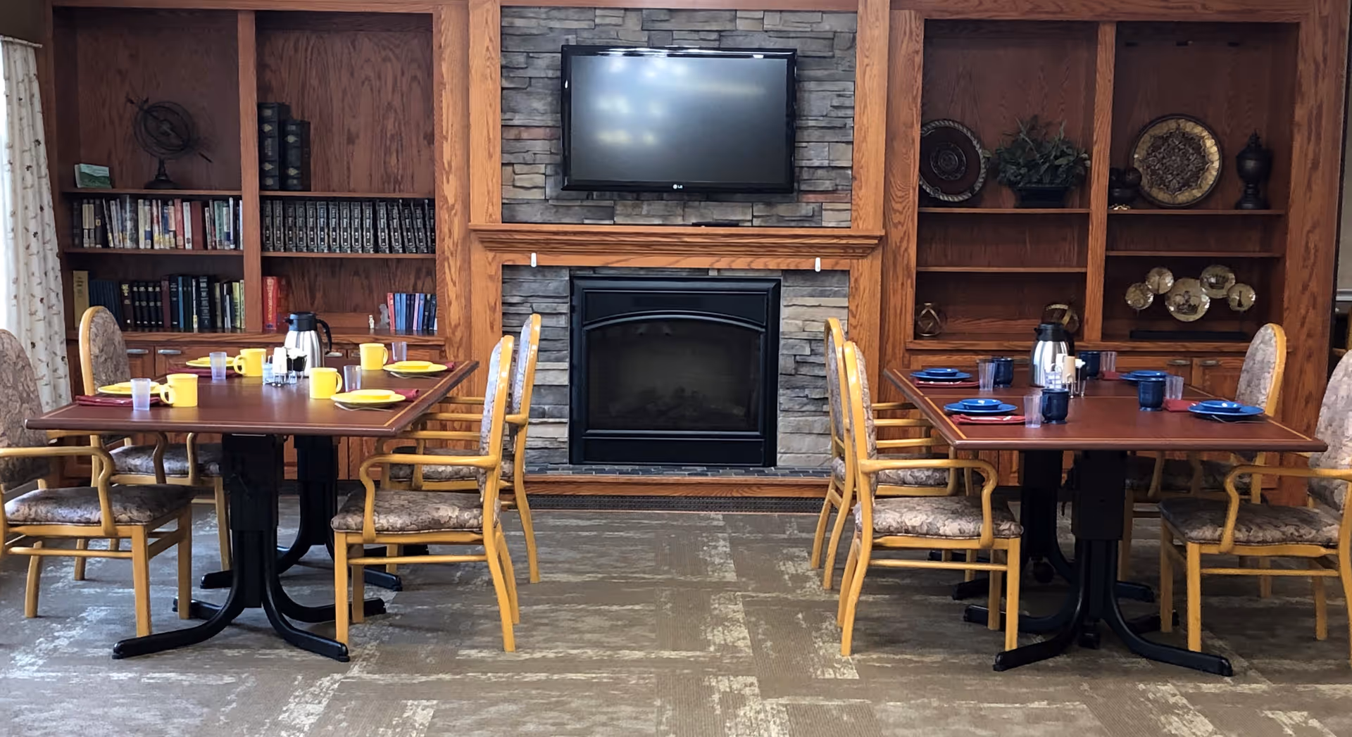 Communal dining area with two set tables and chairs facing a fireplace and wall-mounted TV flanked by wooden bookshelves.