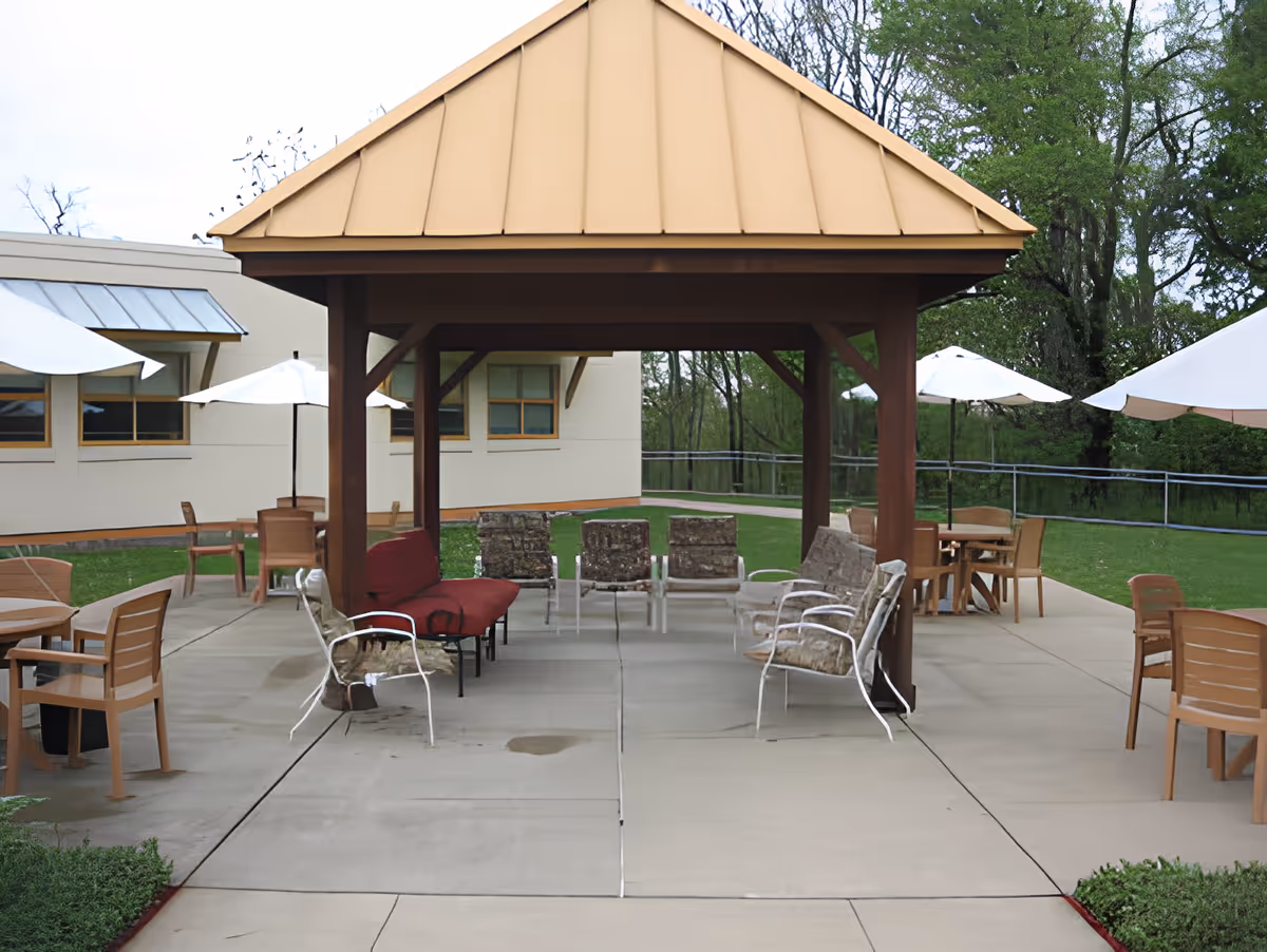 Outdoor seating area with a covered pavilion featuring a tan roof and wooden supports. Several cushioned chairs and a red bench are arranged under the pavilion. Surrounding the pavilion are multiple tables with chairs and white umbrellas on a concrete patio. Green grass and trees are visible in the background.