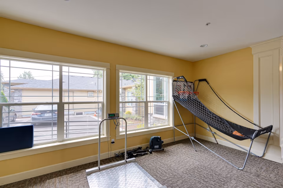 Bright carpeted activity room with large windows, a basketball arcade shooting game, a scale and some exercise equipment against yellow walls.