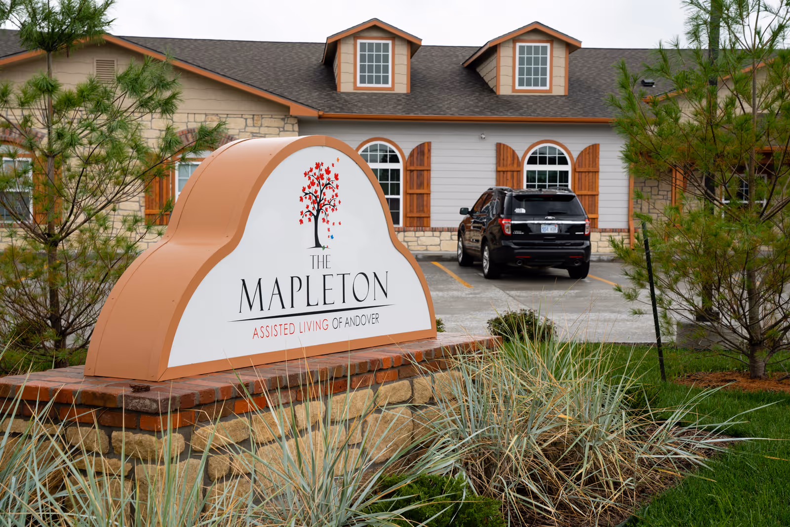 Exterior view of The Mapleton Assisted Living facility with a prominent sign in the foreground displaying the facility's name and logo, a tree with red leaves. The building behind has stone and wood siding, windows with wooden shutters, and a parked black SUV in the parking lot.