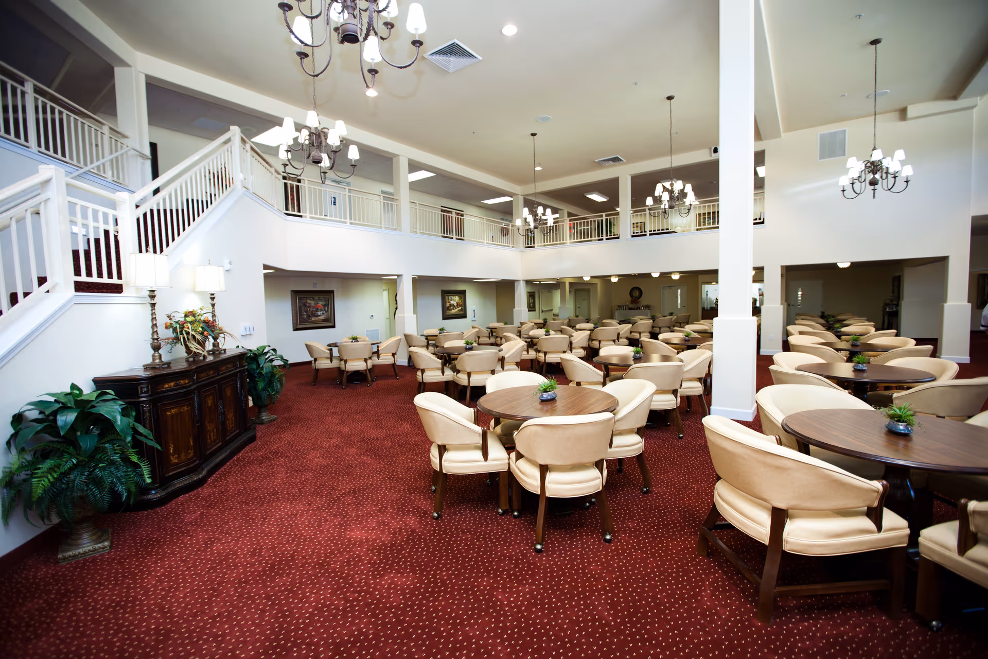 Spacious two-story dining/lounge area with round tables and beige chairs, red carpet, chandeliers and a staircase leading to an upper balcony.