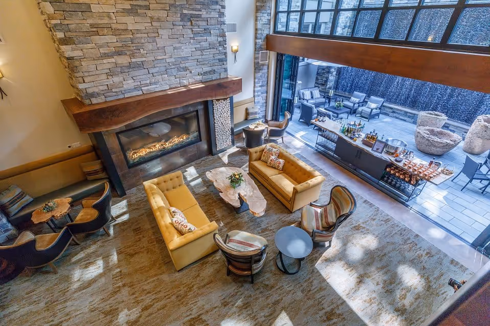 A spacious and well-lit living area with two yellow sofas, two armchairs, and a large stone fireplace. The room features a high ceiling and large windows that open to an outdoor patio with seating and a water feature. A long table with drinks and snacks is set up near the patio entrance.