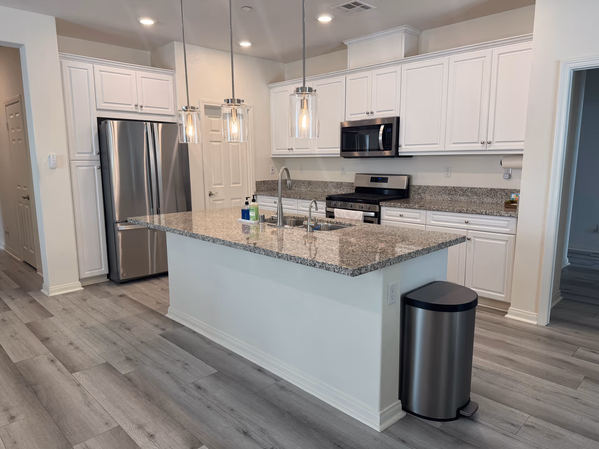 Bright modern kitchen with a large granite island, white cabinetry, stainless steel refrigerator and oven, and pendant pendant lights.