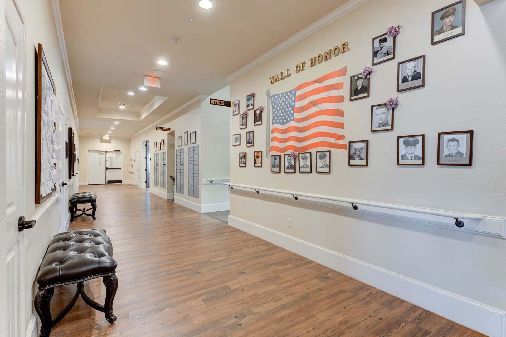 A well-lit hallway in a senior living facility with wooden flooring and white walls. On the right wall, there is a 'Wall of Honor' display featuring an American flag and framed black-and-white photographs of veterans. There are handrails along the wall and two black cushioned benches on the left side. The hallway leads to doors and has signs indicating offices and other rooms.