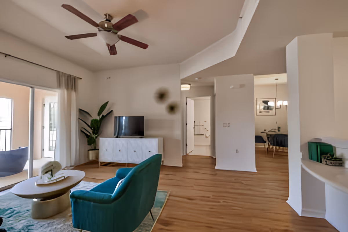 A modern living room with a teal armchair, a round coffee table with a decorative item, a white media console with a flat-screen TV, a large potted plant, and a ceiling fan with wooden blades. The room has wooden flooring and large sliding glass doors leading to a balcony with outdoor seating. In the background, there is a view into a dining area and a hallway leading to other rooms.
