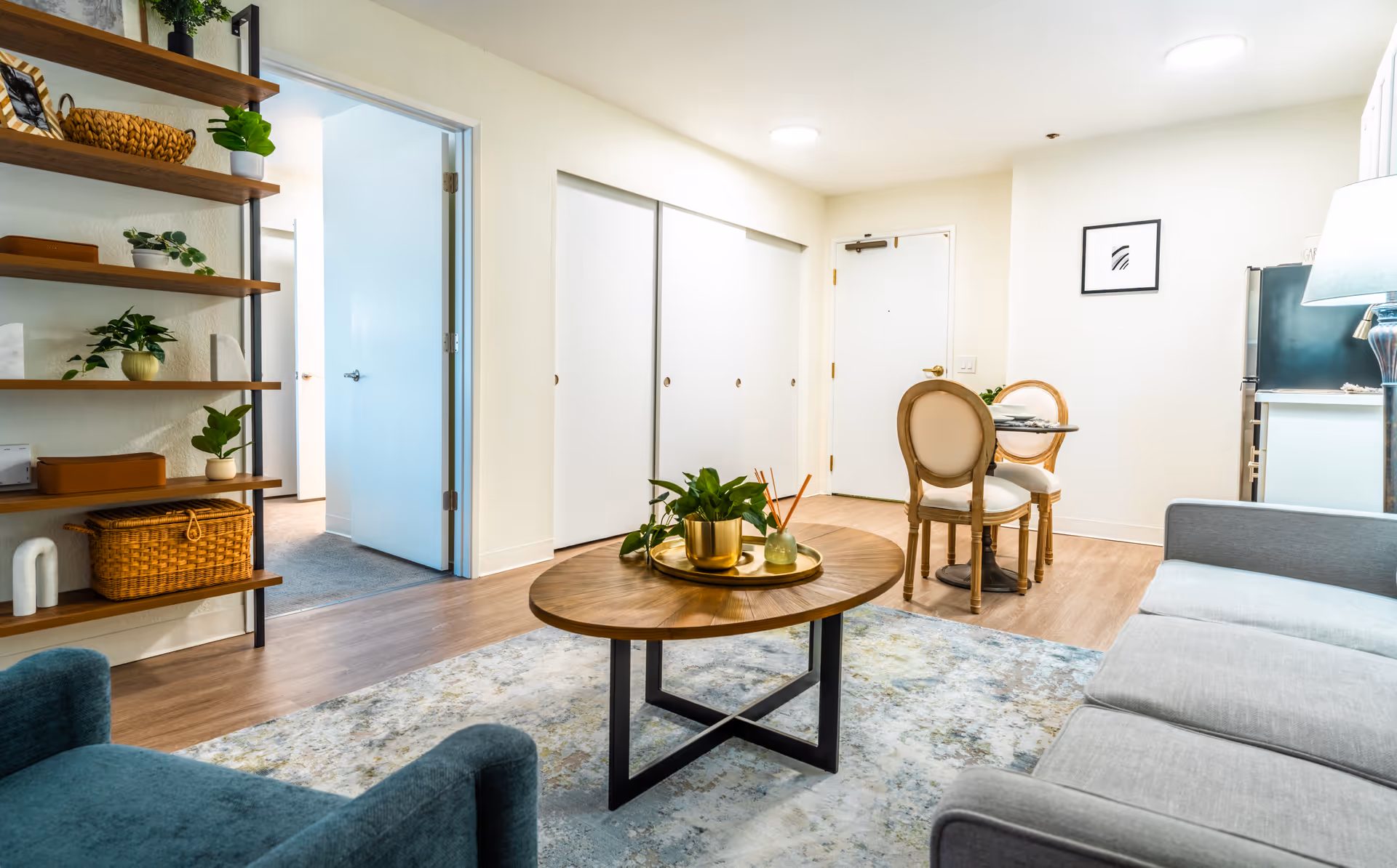 A bright and cozy living room area in a senior living facility featuring a gray sofa, a round wooden coffee table with plants and decorative items, a small dining table with two chairs near the entrance door, wooden shelves with plants and baskets on the left wall, and a light blue armchair in the foreground.