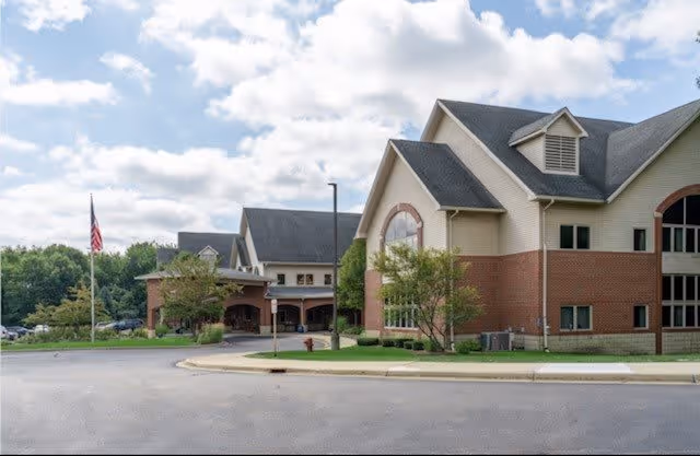 Exterior view of a senior living facility building with a combination of brick and beige siding, large windows, and a sloped roof. There is a driveway in front, a flagpole with an American flag, some trees, and a partly cloudy sky.