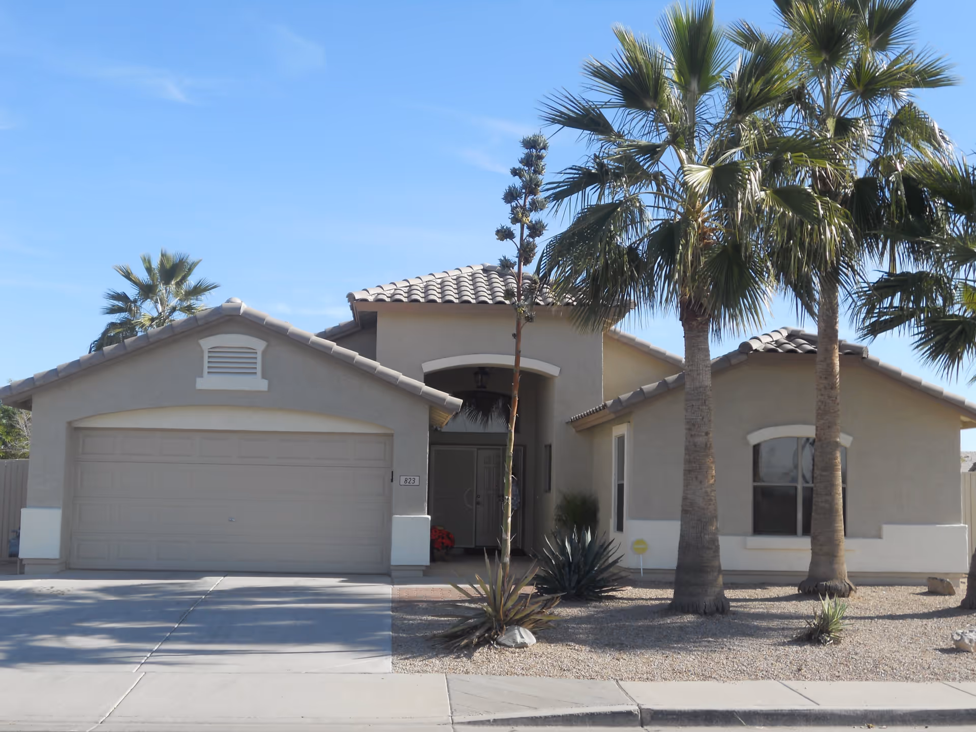 Front exterior of a single-story stucco house with a two-car garage, palm trees, and desert landscaping.