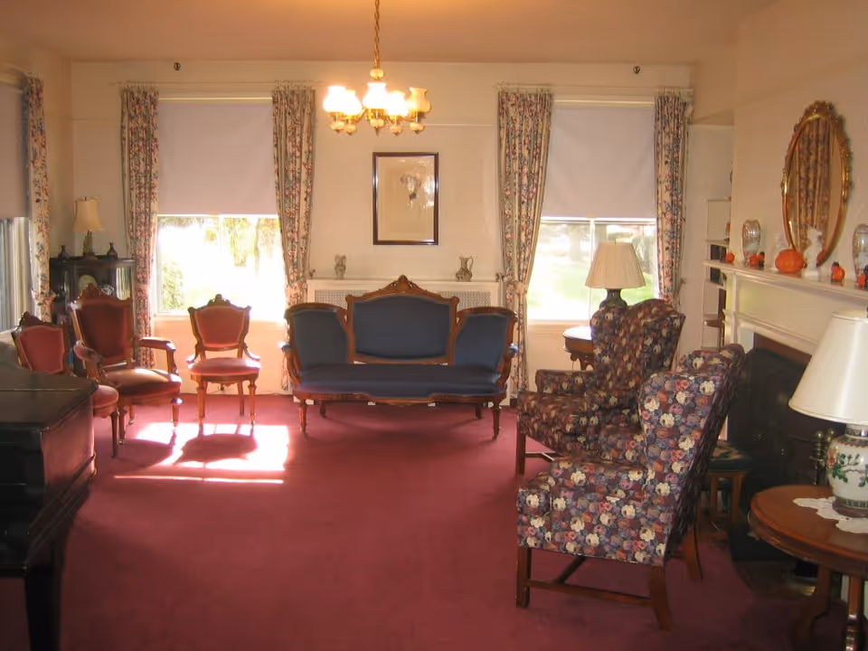 A traditional living room with vintage-style furniture including a blue upholstered sofa, floral armchairs, and wooden chairs with red cushions. The room has floral curtains, a red carpet, a chandelier, and a fireplace with a mirror and decorative items on the mantel.