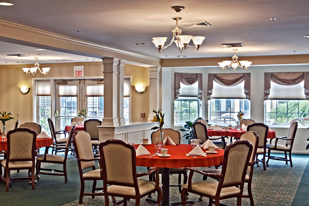 A dining room with round tables covered in red tablecloths, each set with white napkins, cups, glasses, and silverware. The room has upholstered chairs with wooden frames, large windows with draped valances, and ceiling light fixtures. There are plants near the windows and an exit door in the background.
