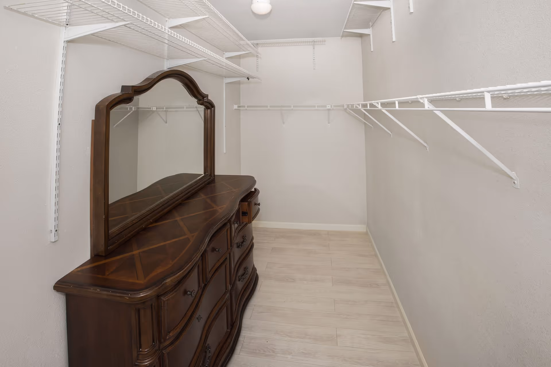 Empty walk-in closet with white wire shelving and a wooden dresser topped by a mirror.
