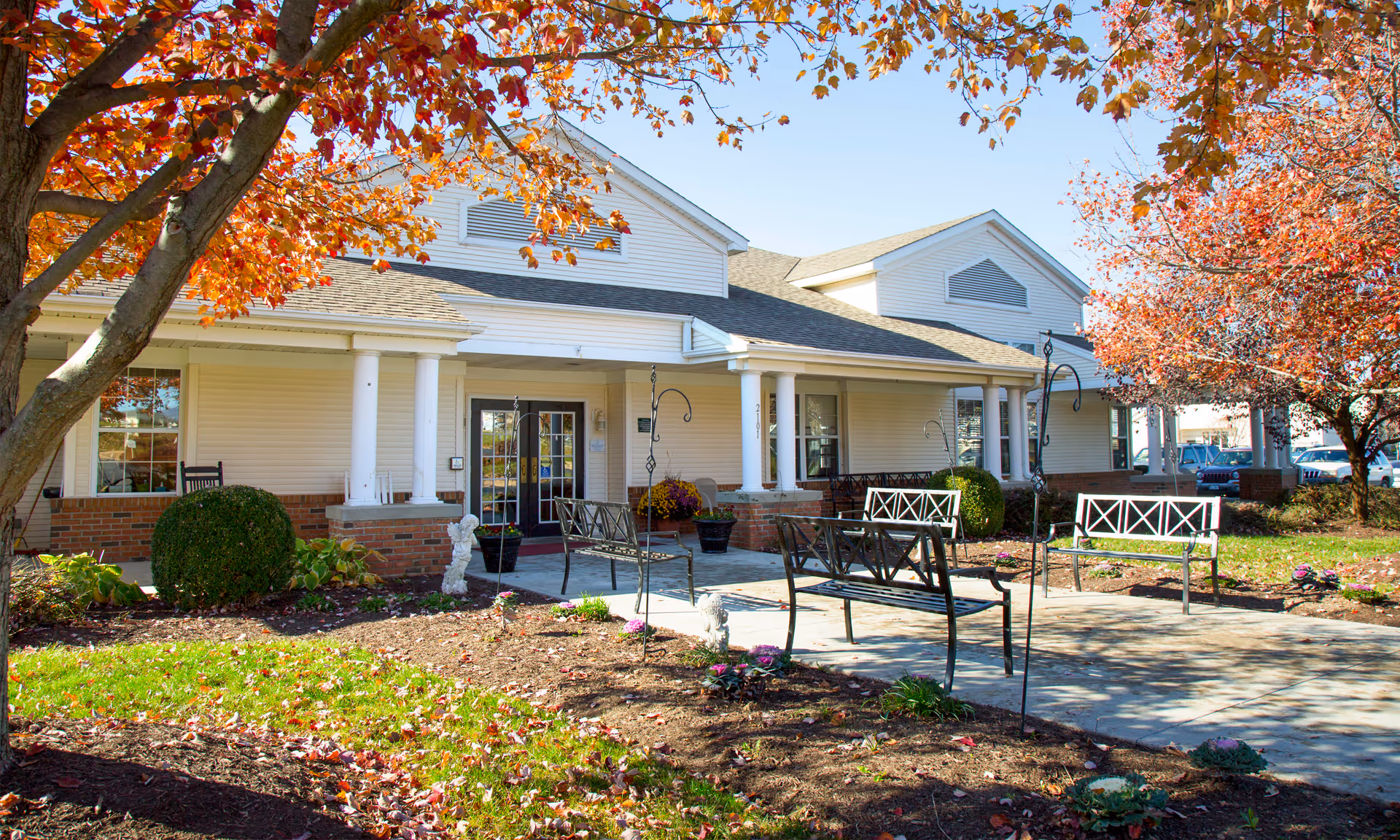 Front entrance of a single-story senior living building with white columns, outdoor benches, and autumn trees.