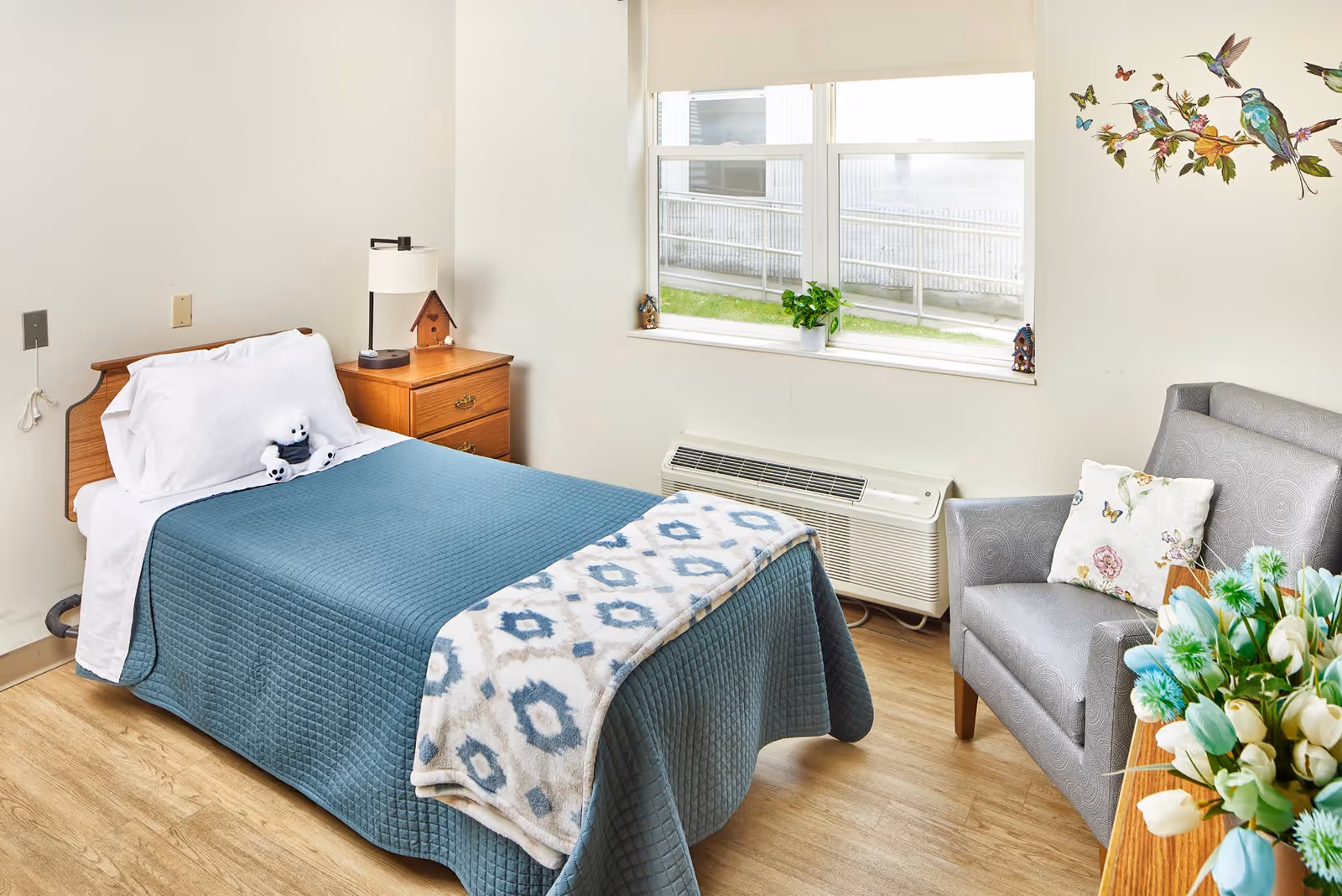 A cozy bedroom featuring a single bed with a blue quilt, a bedside table with a lamp, and a plush panda toy. A comfortable gray armchair sits beside a window with a small plant, and a decorative floral arrangement is on a nearby table.