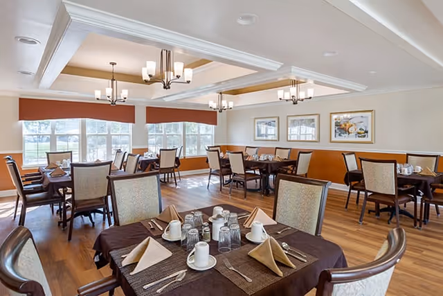 A bright dining room with multiple tables set for meals, featuring brown tablecloths, beige napkins folded in a triangle, white cups, glasses, and silverware. The room has large windows letting in natural light, wooden flooring, and modern chandeliers hanging from a coffered ceiling. Framed artwork decorates the walls.