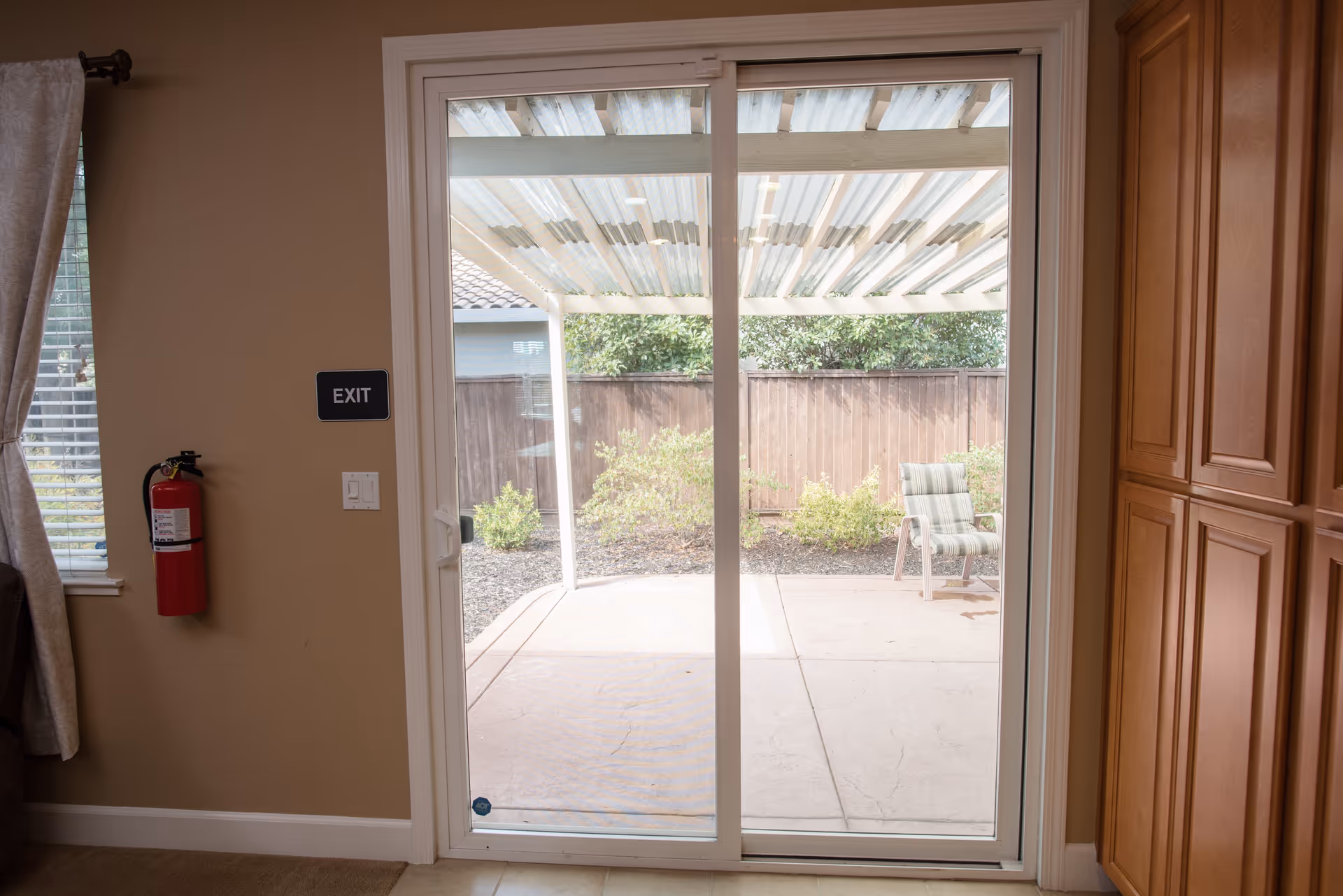 Sliding glass patio door in an interior room looking out to a covered patio with a chair, an EXIT sign and a fire extinguisher on the wall.
