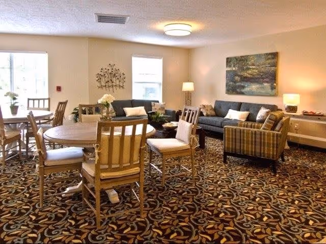 Well-lit communal living room with sofas, armchairs, round dining tables and a patterned carpet.