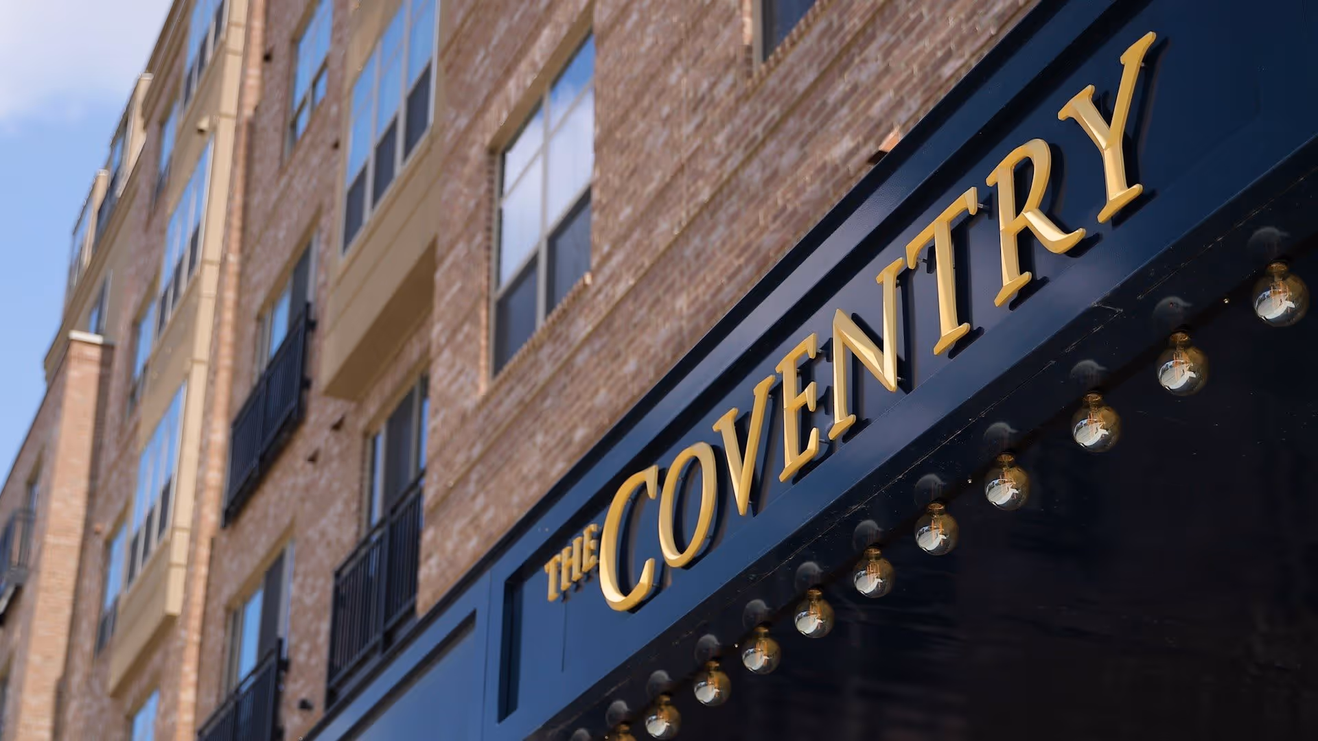 Exterior view of a multi-story brick building with large windows and balconies, featuring a sign that reads 'The Coventry' in gold letters on a dark blue background with light bulbs underneath.