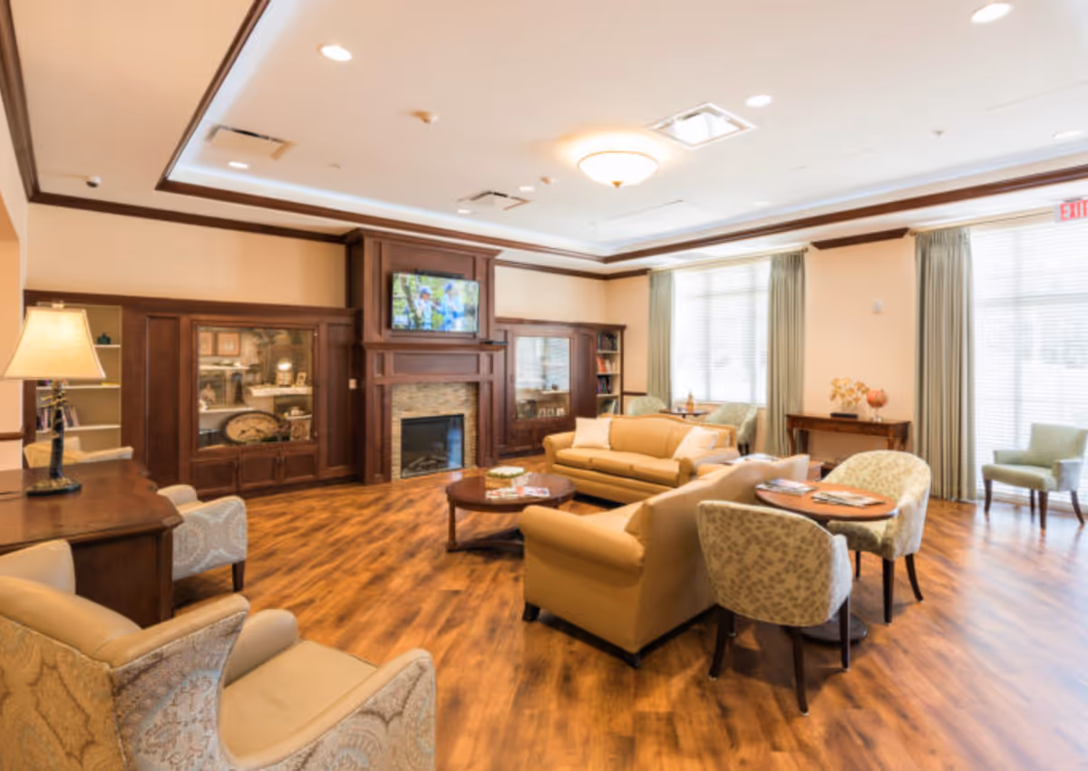 A spacious and well-lit living room with wooden flooring, beige sofas, patterned armchairs, a round wooden coffee table, and a fireplace with a mounted flat-screen TV above it. Large windows with curtains allow natural light to fill the room, and there are additional small tables and chairs near the windows.