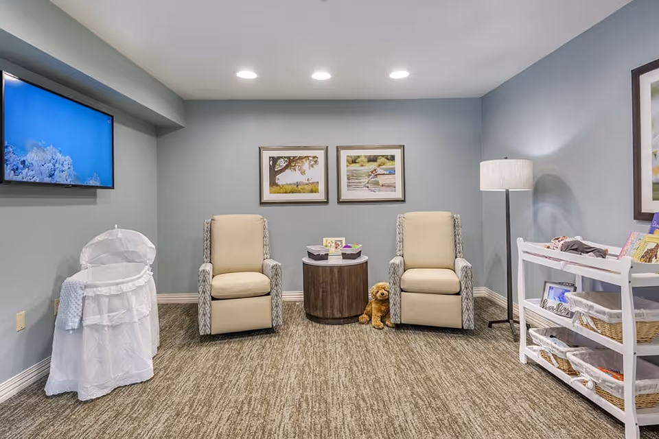A cozy room with two beige armchairs with patterned sides, a round wooden table between them holding baskets and small items, a stuffed dog toy on the floor, a white bassinet on the left, a white shelving unit with baskets and books on the right, a floor lamp, two framed pictures on the wall, and a flat-screen TV mounted on the left wall.