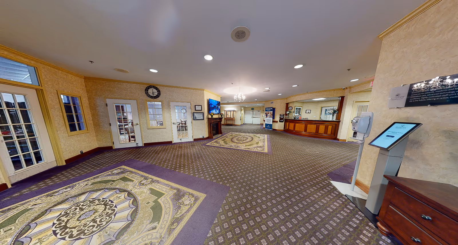 Spacious carpeted reception lobby with decorative rugs, a front desk, entry doors, and a kiosk.