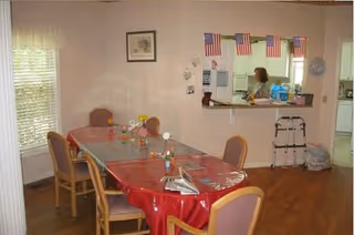 Dining room with a long table covered in red plastic and chairs, American flags above a kitchen pass-through with a person standing behind it.