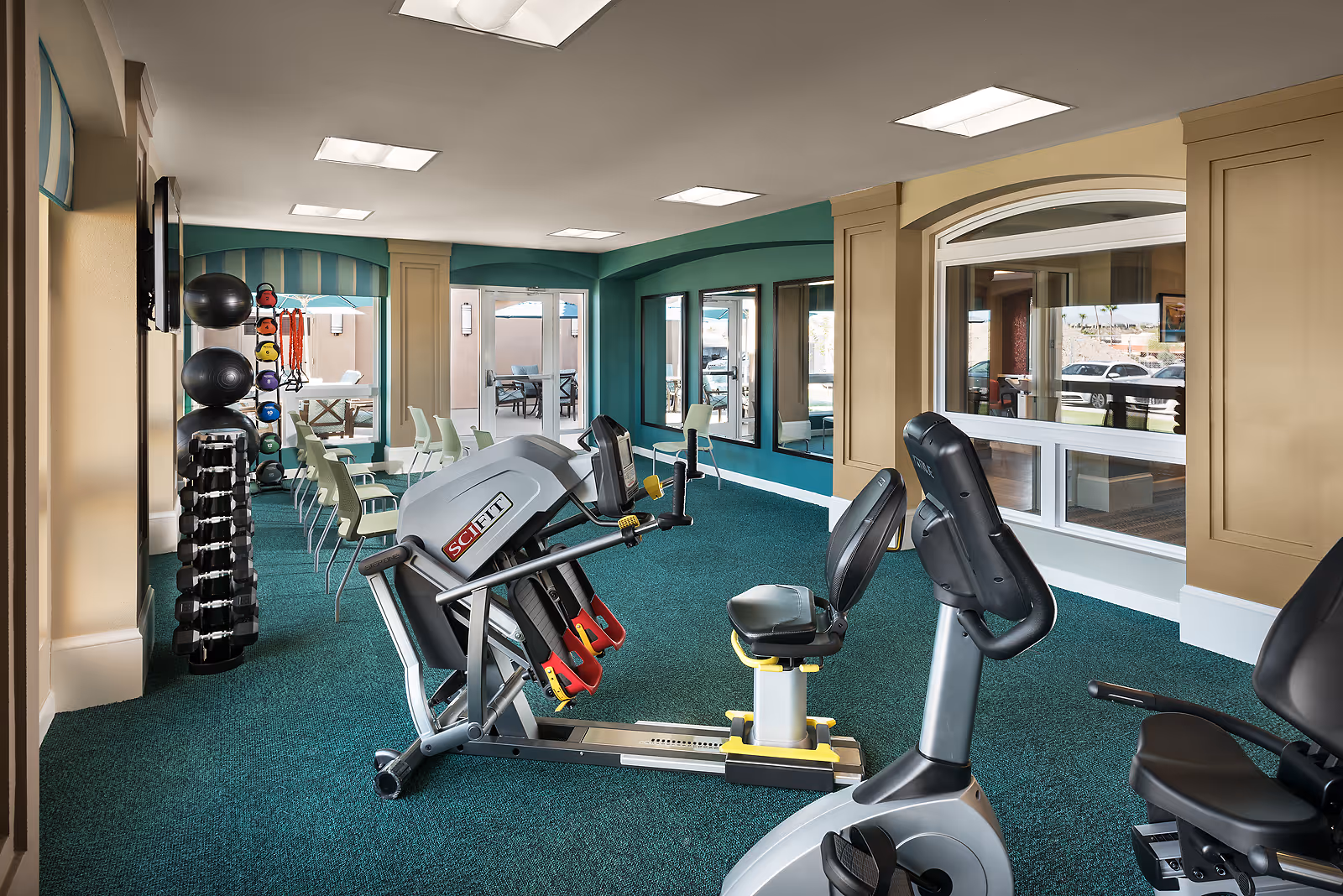 Interior view of a fitness room in MorningStar Assisted Living & Memory Care of Fountain Hills featuring exercise equipment including a recumbent bike and a SciFit machine, a rack of dumbbells and medicine balls, chairs lined up along the wall, and large windows with a view of an outdoor patio area.