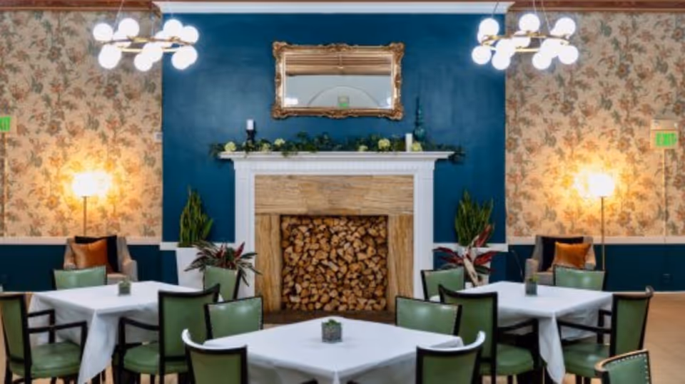 A cozy dining area with four tables covered in white tablecloths, each surrounded by green chairs. The room features a decorative fireplace filled with stacked firewood, a large ornate mirror above it, floral wallpaper, and two standing lamps on either side. There are also plants and comfortable armchairs in the corners, with modern ceiling lights illuminating the space.