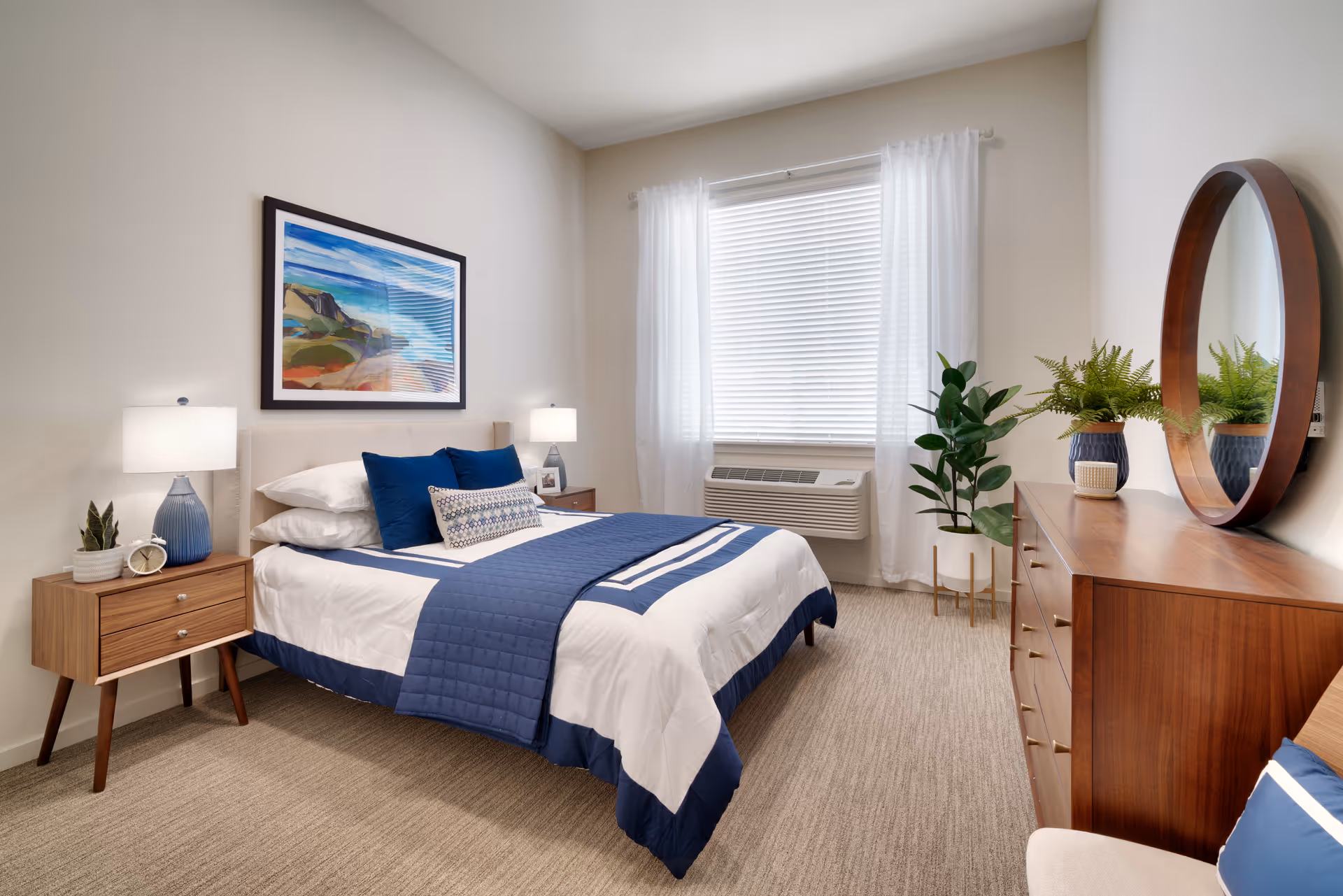 A neatly made bed with white and blue bedding in a bright bedroom. The room features two wooden nightstands with lamps, a framed landscape painting above the bed, a large window with white curtains, a wooden dresser with a round mirror, and two potted plants.