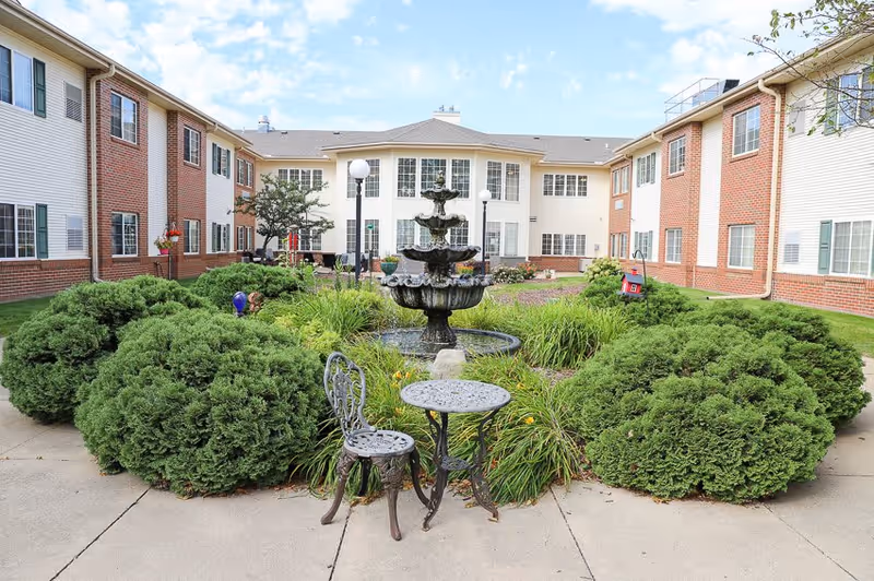 Outdoor courtyard area at The Heritage at Northern Hills featuring a three-tiered water fountain surrounded by green bushes and plants. There is a small metal table with two matching chairs on a concrete pathway. The courtyard is enclosed by a two-story building with white siding and red brick accents under a partly cloudy sky.