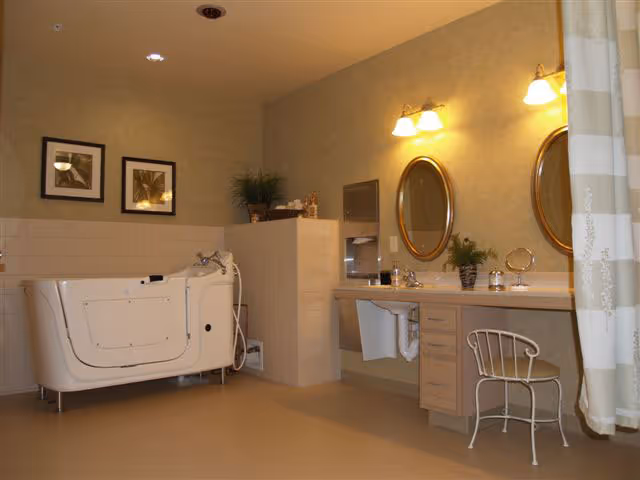 A spacious bathroom featuring a walk-in bathtub with safety handles, a vanity area with two oval mirrors and wall-mounted lights above, a small chair, and decorative plants. The walls are tiled halfway up and painted beige above, with framed pictures hanging on the wall.