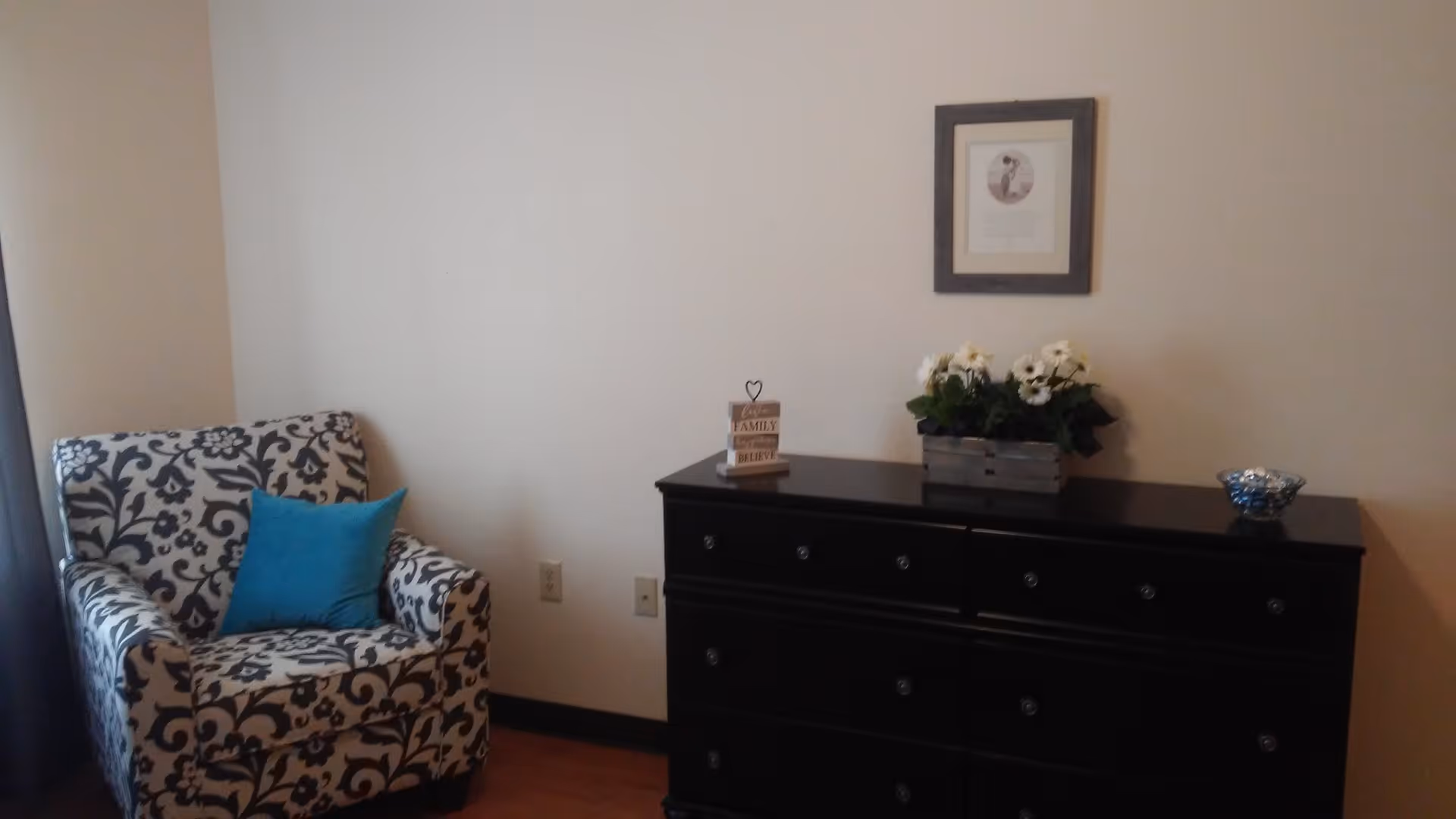 A cozy corner of a room featuring a patterned armchair with a blue cushion on the left and a black dresser on the right. On top of the dresser are a small decorative sign, a planter with white flowers, and a small bowl. A framed picture hangs on the wall above the dresser.