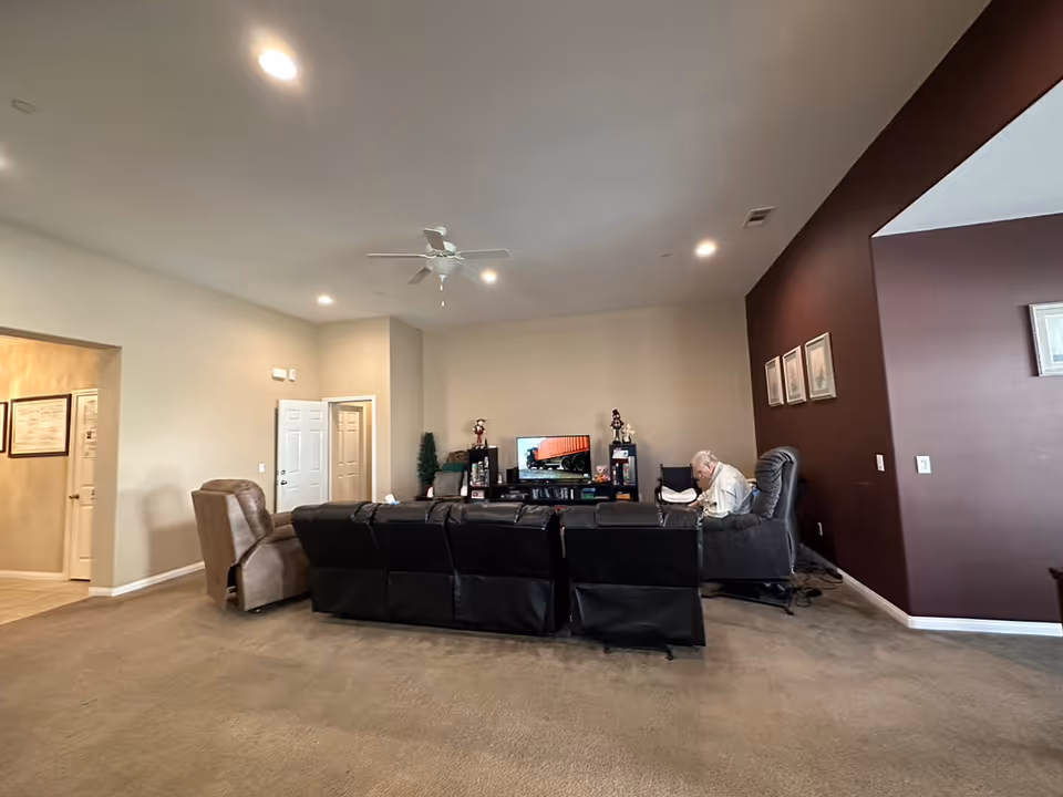 A spacious living room in an assisted living facility with beige walls and carpeted floor. The room features a black leather sectional sofa and a brown recliner chair. A television is placed on a black entertainment unit against the far wall, with decorative items and framed pictures on the walls. An elderly person is seated on the right side of the sofa, engaged in an activity. The ceiling has recessed lighting and a white ceiling fan.