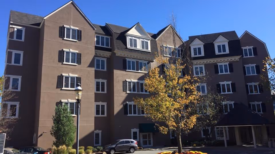 Front view of a multi-story brown rehabilitation and skilled nursing building with many windows, a tree and parked cars under a clear blue sky.