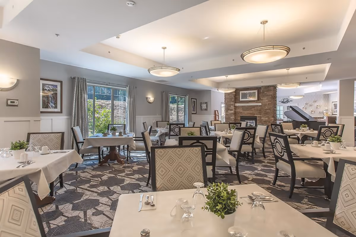 A bright and spacious dining room with multiple tables covered in white tablecloths, each set with glasses, silverware, and small green plants. The room features large windows with curtains allowing natural light to enter, a stone fireplace, and a grand piano in the background. The ceiling has modern circular light fixtures.