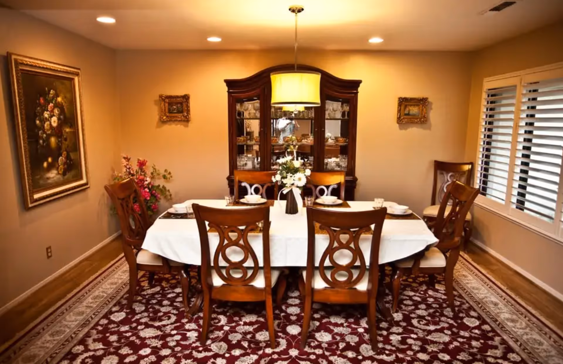 A formal dining room with a rectangular table covered with a white tablecloth, set for six people with plates, glasses, and napkins. The room features wooden chairs with decorative backs, a floral centerpiece on the table, a large wooden china cabinet filled with glassware and dishes, a hanging light fixture above the table, a patterned red and white rug, a large floral painting on one wall, two smaller framed pictures on the opposite wall, and a window with white blinds.