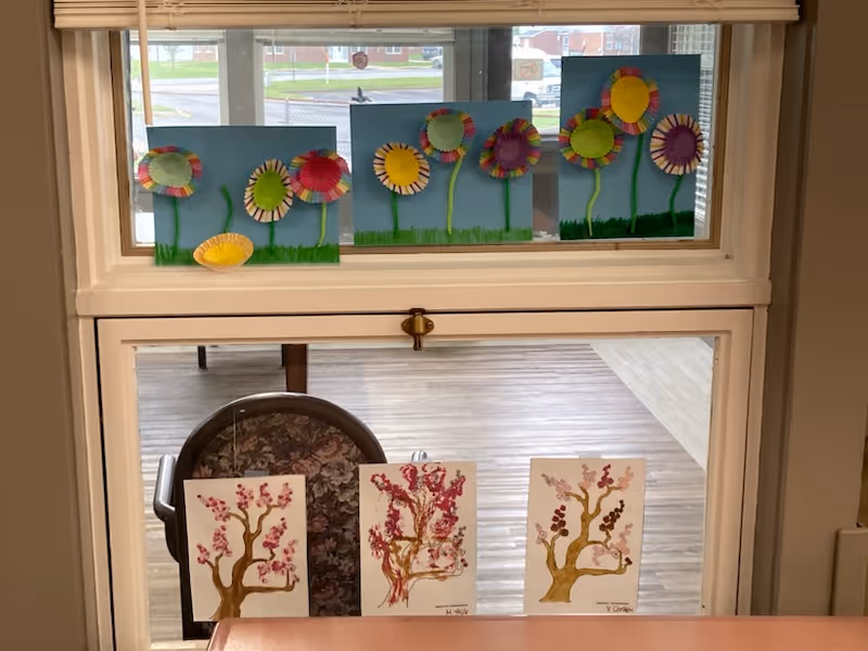 A window with colorful handmade flower crafts displayed on the upper pane, made from paper plates and pipe cleaners on blue paper backgrounds. Below, three paintings of blossoming trees are placed on a table in front of a floral-patterned chair. The room has wooden flooring and a bright, airy atmosphere.