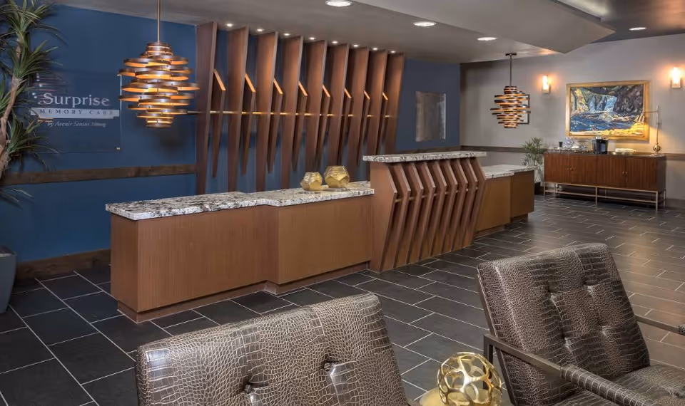 Reception area of Mosaic Gardens Memory Care at Surprise featuring a modern wooden front desk with a granite countertop, two pendant lights hanging above, a blue accent wall with the facility's name, and comfortable seating with patterned chairs in the foreground. There is also a sideboard with a coffee station and a framed painting on the wall.