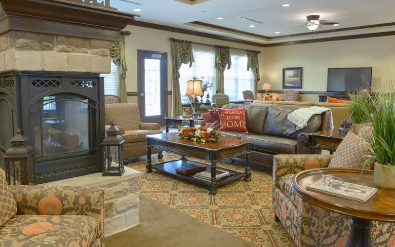 A cozy living room featuring a stone fireplace, comfortable seating, and decorative accents. The room is well-lit with natural light from large windows, creating a warm and inviting atmosphere.