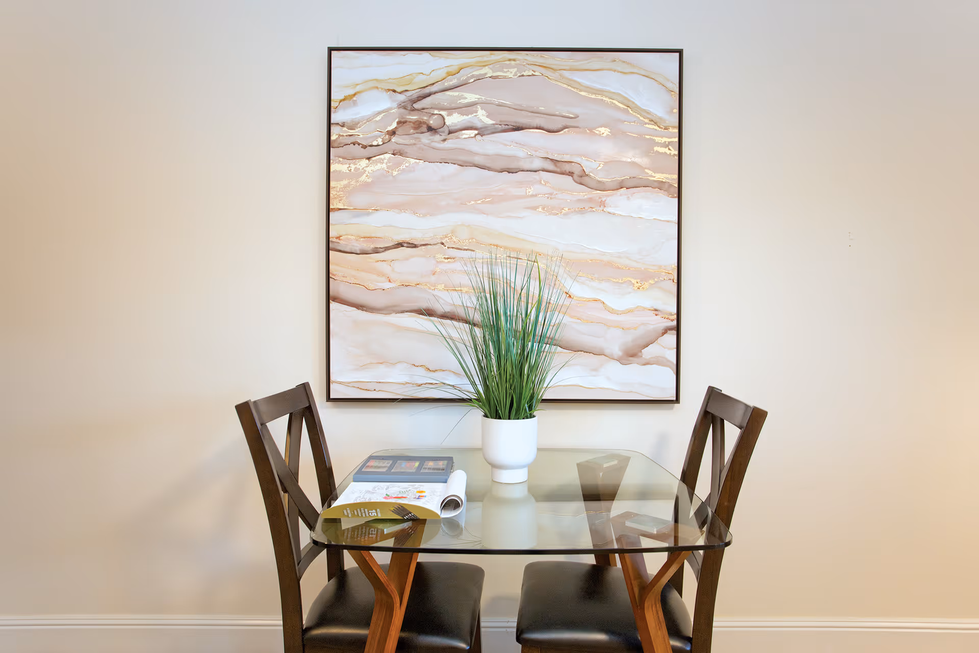 Small dining area with a glass table, two wooden chairs, a potted plant and abstract wall art on the wall.