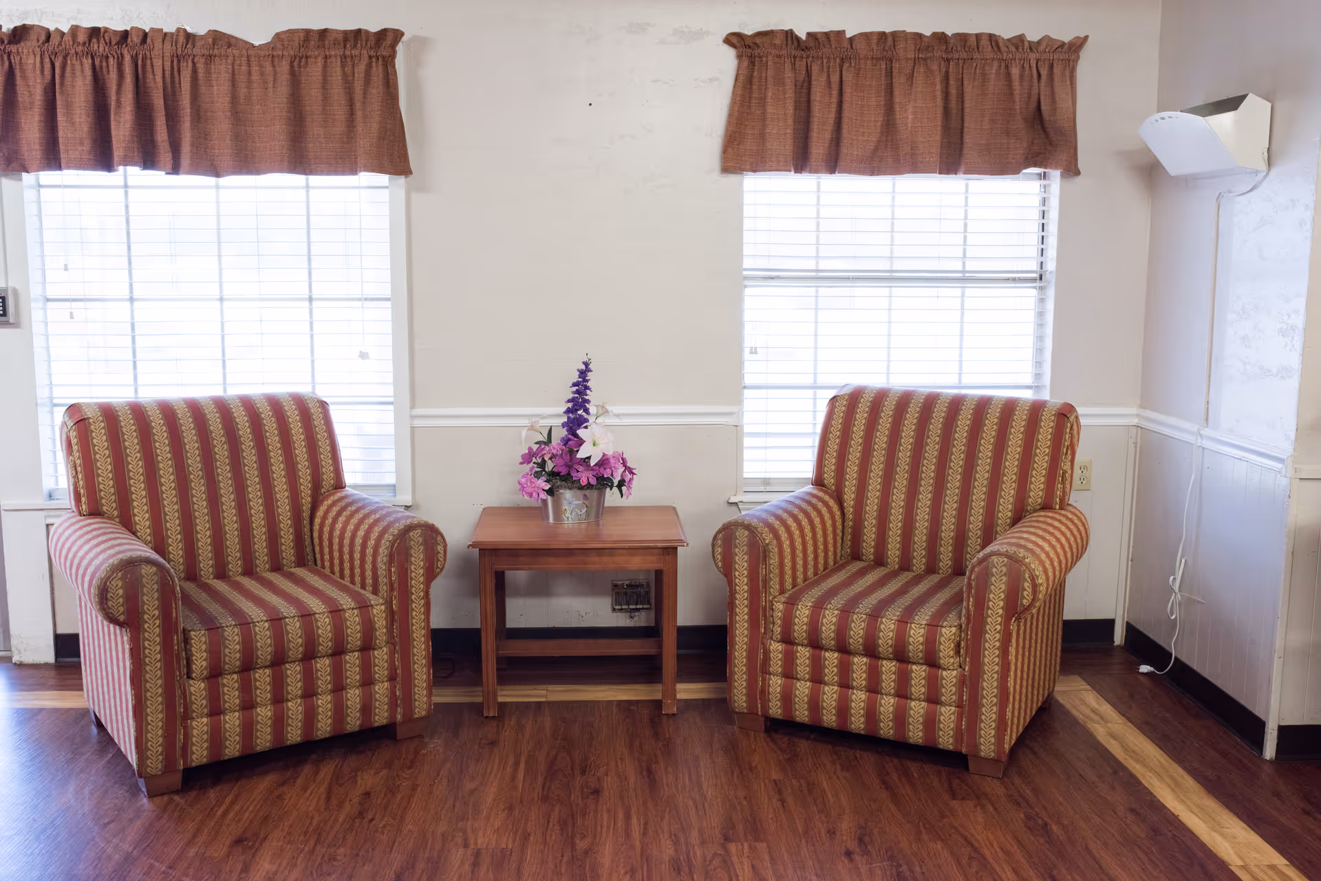 Two striped armchairs with red and gold patterns are placed on either side of a wooden side table with a flower arrangement in a pot. Behind them are two windows with white blinds and brown valance curtains. The room has wooden flooring and light-colored walls with white wainscoting.
