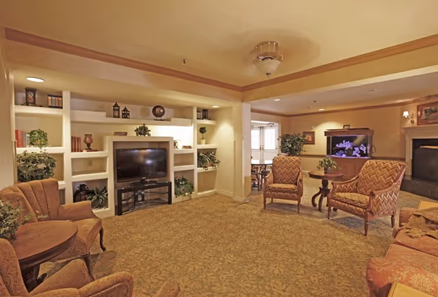 A cozy living room area in a senior living facility featuring patterned armchairs and sofas arranged around a wooden coffee table. There is a built-in white shelving unit with decorative items and a flat-screen TV. A fish tank is visible near a fireplace, and the room is warmly lit with ceiling lights and wall sconces.