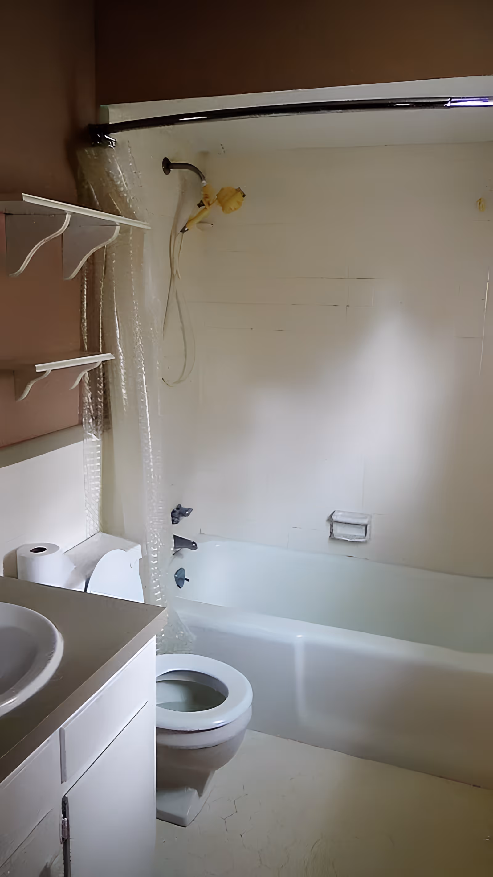 A small bathroom with a white bathtub and shower combination, a white toilet, a white sink with a countertop, and two white wall shelves. The shower has a curved shower rod with a clear shower curtain. The walls are painted brown and white.